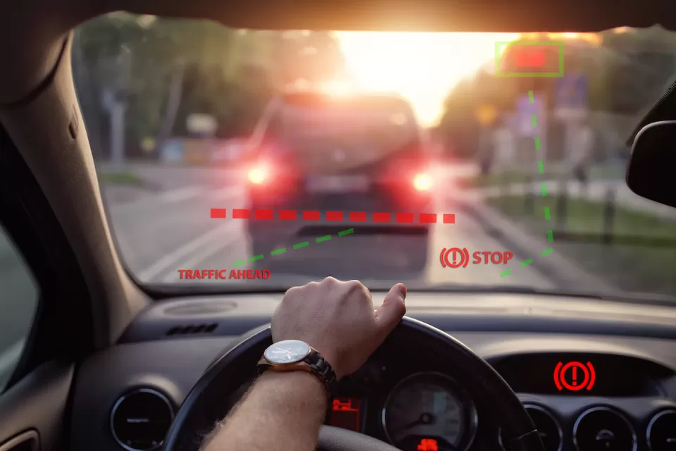 Close-up of car steering wheel and dashboard with driver's hand on steering wheel and illustration of automatic braking system on windshield with 'STOP' graphic.